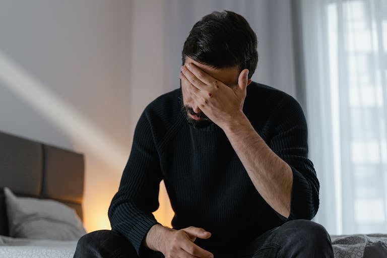 A man in distress sits on a bed indoors, covering his face with his hand.
